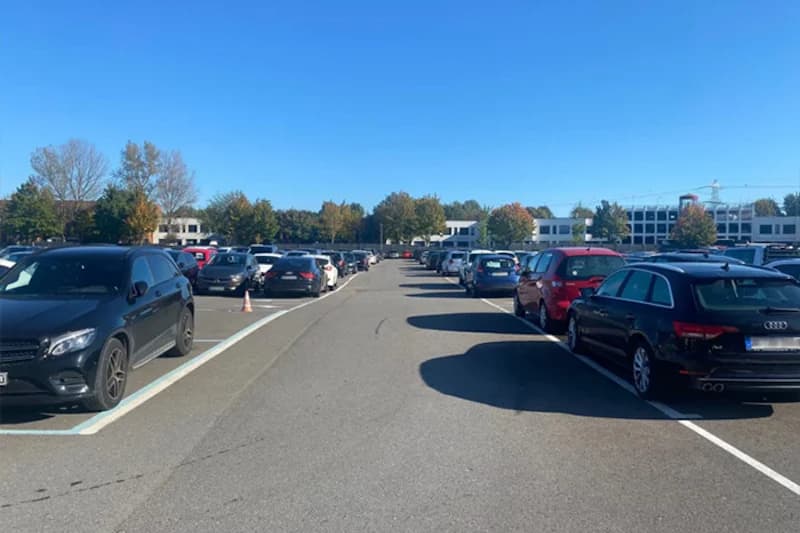 Großer Außenparkplatz mit zwei Reihen geparkter Autos, Bäumen und Gebäuden im Hintergrund.