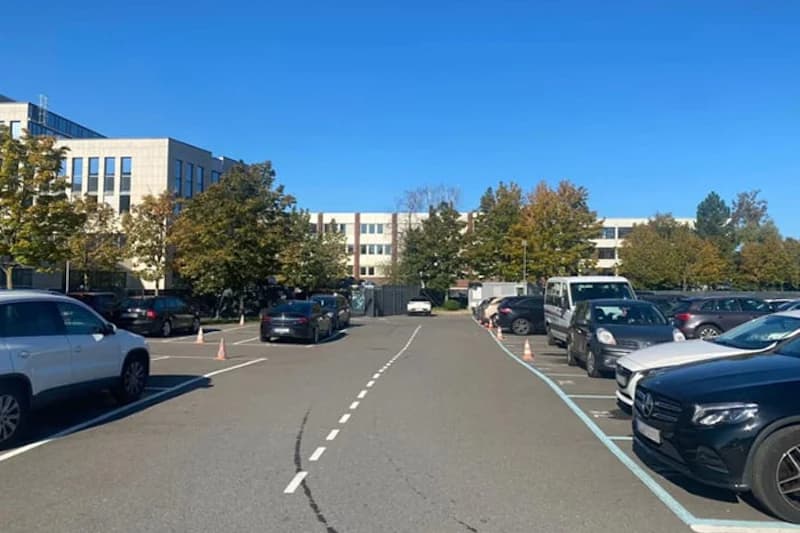 Parkplatz mit geparkten Autos links und rechts, Zaun und Bürogebäude im Hintergrund.