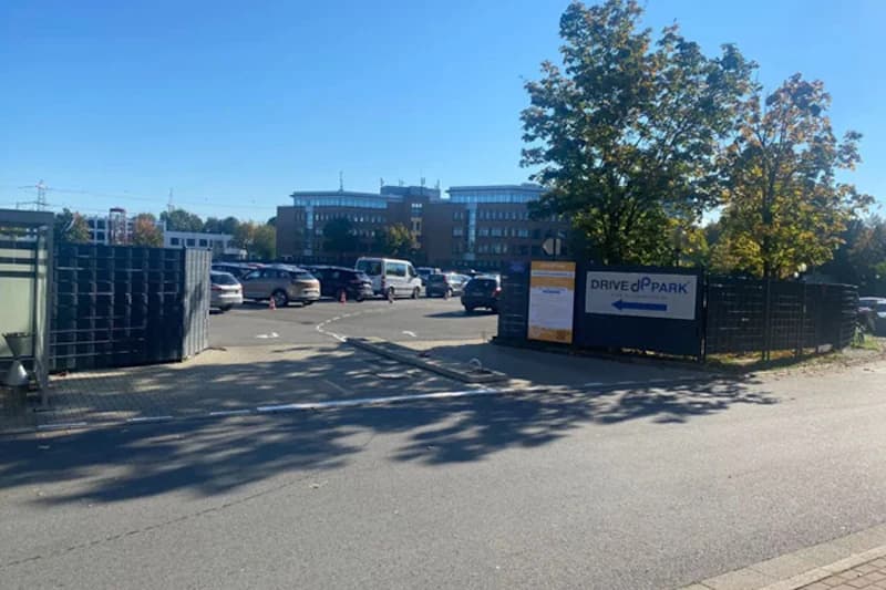 Einfahrt zu einem umzäunten Parkplatz mit geöffnetem Tor, Drive&Park-Schild, geparkten Autos und Bürogebäude im Hintergrund.