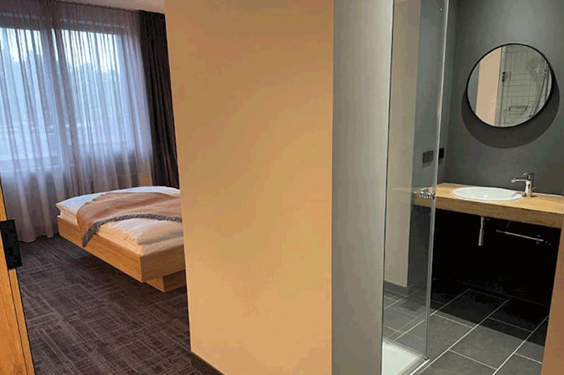Hotelzimmer mit Bett am Fenster, halb offenes Bad mit Glasdusche, Waschbecken auf Holzplatte und rundem Spiegel.