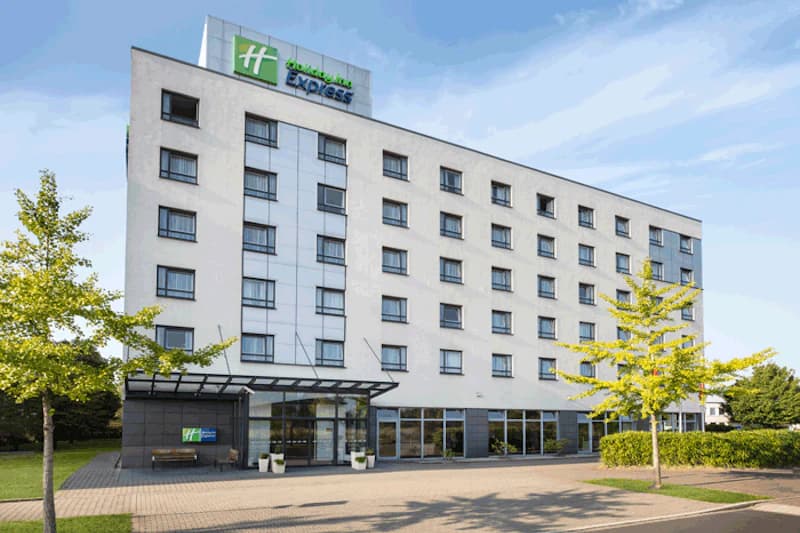 Fünfgeschossiges Hotelgebäude mit Glasvordach, Holiday Inn Express Schild auf dem Dach und gepflasterter Zufahrt mit Bäumen.