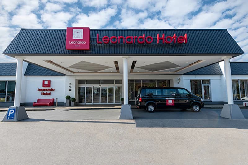 Eingang des Leonardo Hotel mit Vordach, rotem Schriftzug, geparktem Hotelbus und Parkplatzschild.