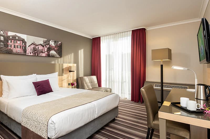Hotelzimmer mit Doppelbett, Kopfkissen, Lesesessel, Schreibtisch mit Lampen und Teekocher sowie Fenster mit Vorhängen.