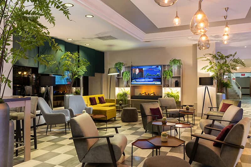 Hotel-Lounge mit Sesseln, Sofas, Beistelltischen, Fernseher über elektrischem Kamin und großen Zimmerpflanzen.