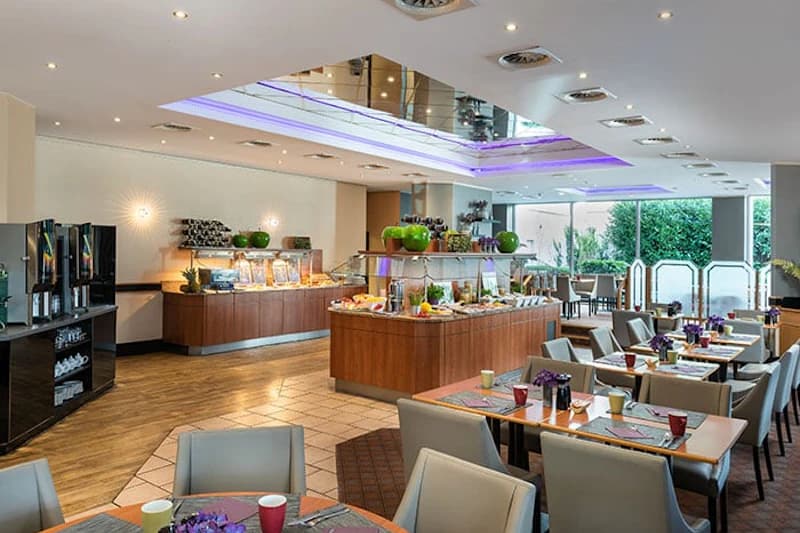 Großer Hotelfrühstücksraum mit Buffetinsel, gedeckten Tischen und großen Fenstern zur Gartenansicht.