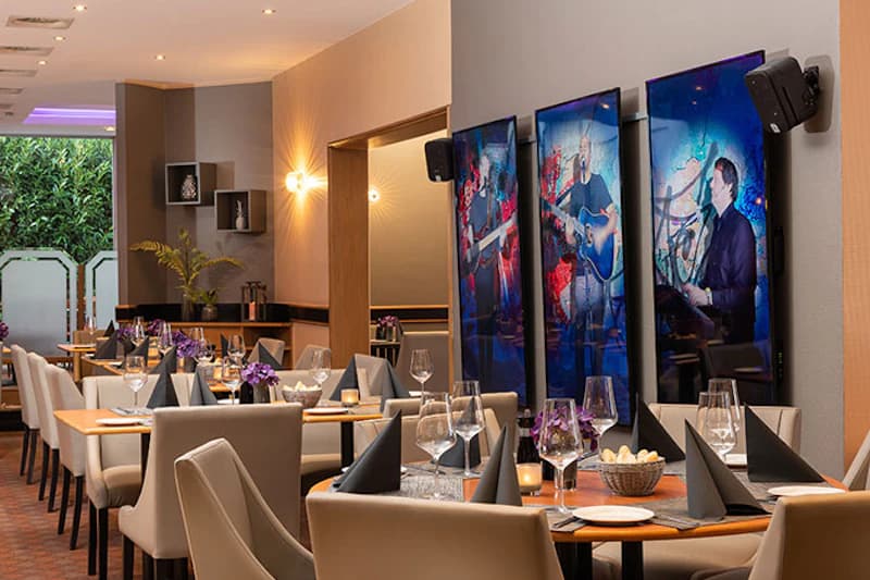 Gedeckte Tische in Hotelrestaurant mit beigen Stühlen, schwarzen Servietten und drei Bildschirmen mit Musikern.