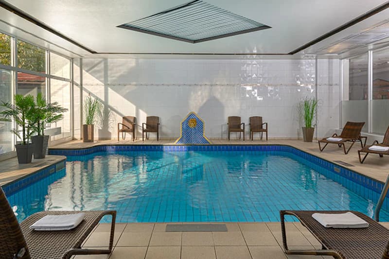 Innenpool mit blau gefliestem Becken, Liegestühlen, Pflanzen und Oberlicht.