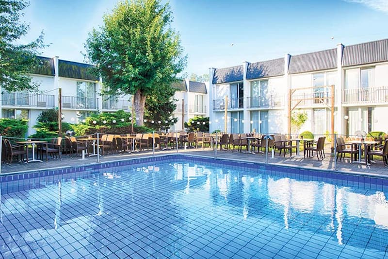 Außenpool mit blau gekacheltem Becken, Terrassen mit Tischen und Stühlen vor zweigeschossigem Hotelgebäude und Bäumen.