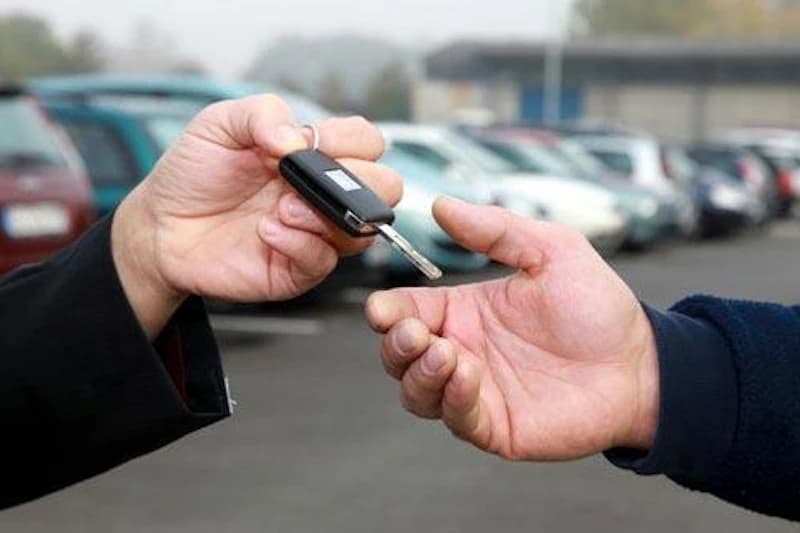 Zwei Hände reichen einen Autoschlüssel vor geparkten Autos auf einem Parkplatz.