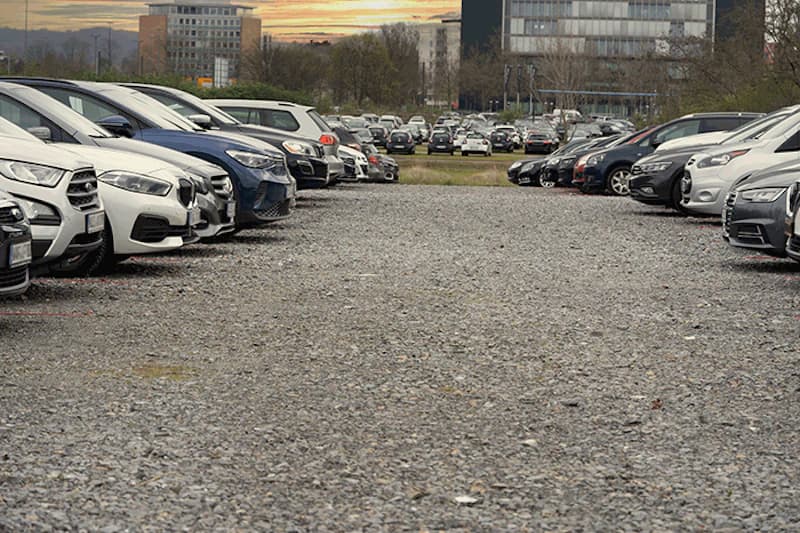 Schotterparkplatz mit zwei Reihen geparkter Autos und Bürogebäuden im Hintergrund bei Sonnenuntergang.