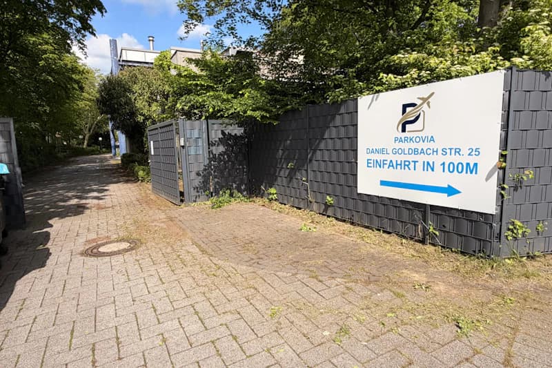 Gepflasterter Zufahrtsweg neben grauem Sichtschutzzaun mit Parkovia-Schild und blauem Pfeil, Bäume im Hintergrund.