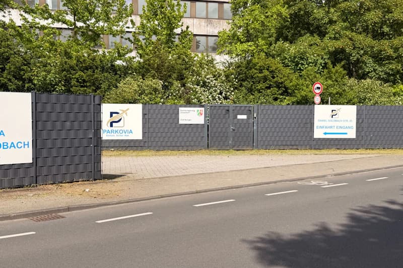Grau eingefasste Zaunfront mit geschlossenem Tor, Parkovia-Logo und Richtungsschild, Bäume hinter dem Zaun, Gehweg und Straße im Vordergrund.