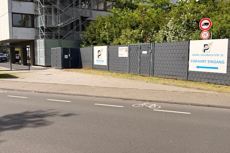 Gesperrte Zufahrt mit hohem Zaun, Parkovia-Schildern, Verkehrszeichen und Fahrradpiktogramm auf der Fahrbahn.