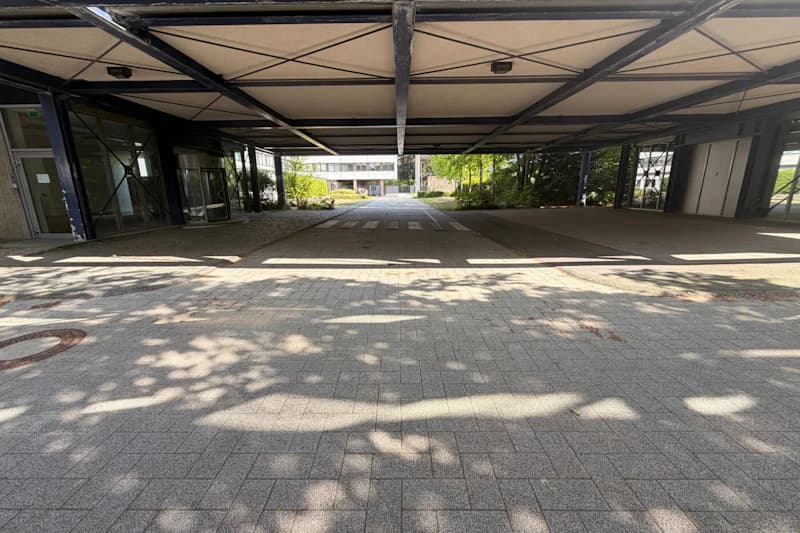 Überdachte Zufahrt vor Gebäude mit Zebrastreifen und Schatten auf dem gepflasterten Boden.