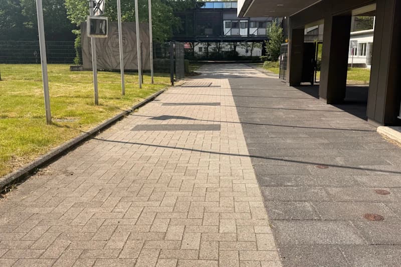 Gepflasterter Fußweg zur geschlossenen Einfahrt mit Metallzaun links und überdachtem Gebäude rechts.