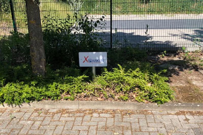 Kleines Metallschild in einem Beet vor Maschendrahtzaun zwischen Baum und Bodendeckern am Parkplatzrand.