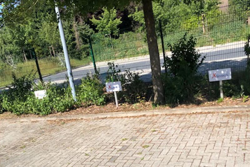 Parkplatzfläche mit drei kleinen Reserviert-Schildern an einem Zaun und einem Baum.