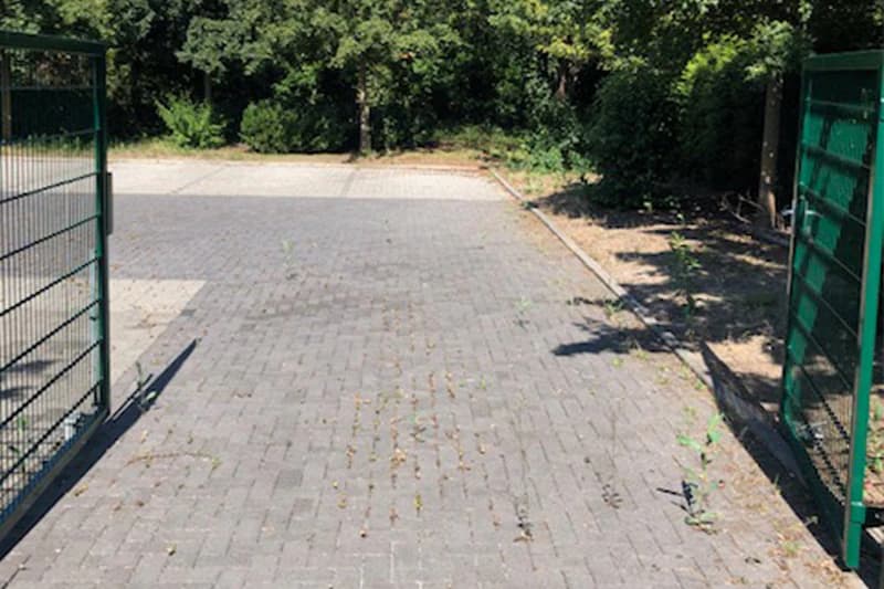 Offener grüner Metallzaun links und rechts, Blick auf gepflasterte Fläche mit Unkraut und Baumbestand im Hintergrund.
