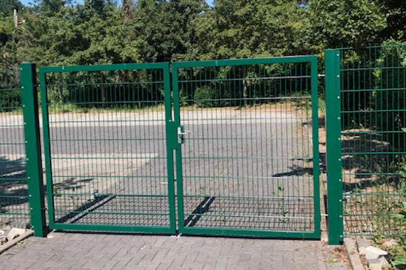 Grünes Doppeltor aus Metallgitter vor asphaltiertem Bereich und Bäumen.