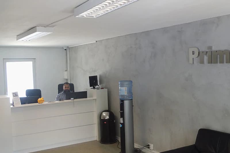 Empfangstheke mit Mitarbeiter am Bildschirm, Trinkwasserspender, Mülleimer und Sofa vor grauer Wand mit Primus-Schriftzug.