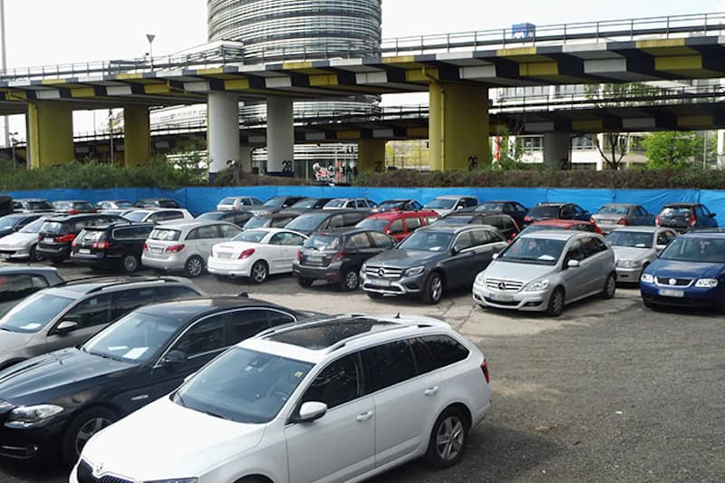 Offener Parkplatz mit dicht gereihten Autos vor erhöhter Fahrbahn auf gelb-schwarzen Stützpfeilern.