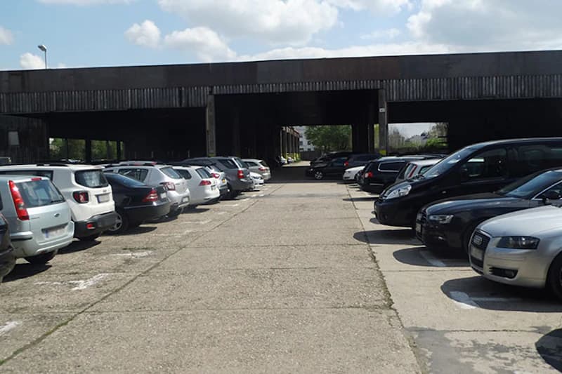 Freier Mittelweg zwischen Reihen geparkter Autos auf offenem Parkplatz mit überdachtem Stahlbau im Hintergrund.