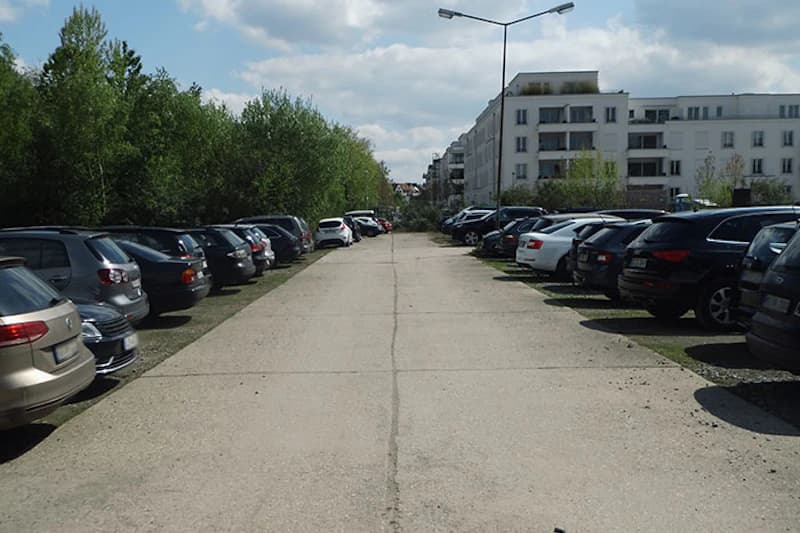 Breite Zufahrt mit geparkten Autos links und rechts, Laterne in der Mitte, Wohnhäuser rechts und Bäume links.