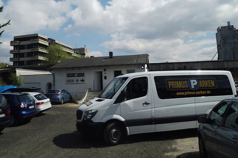 Weißer Primus Parken Transporter auf Schotterplatz vor kleinem Gebäude mit Check-in Schild, mehrere geparkte Autos und bewölkter Himmel.