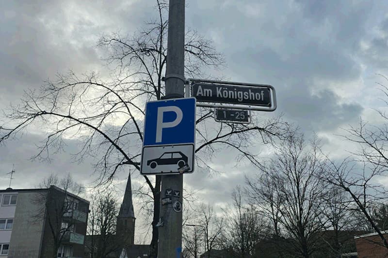 Straßenschild Am Königshof und blaues Parkzeichen mit Autosymbol an Laterne vor bewölktem Himmel.