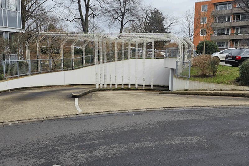 Schräge Zufahrt zur Tiefgarageneinfahrt mit weißem Metallgitter, Geländer und gebogener Auffahrt, Wohngebäude im Hintergrund.