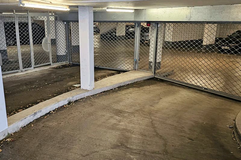 Beleuchtete Auffahrt in Tiefgarage mit Betonrampe, Stützsäule und geschlossenen Metallgittern, geparkte Autos dahinter.