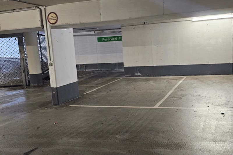 Leere Tiefgaragenstellplätze mit Betonsäulen, Bodenmarkierungen und geschlossenem Metallgittertor links.