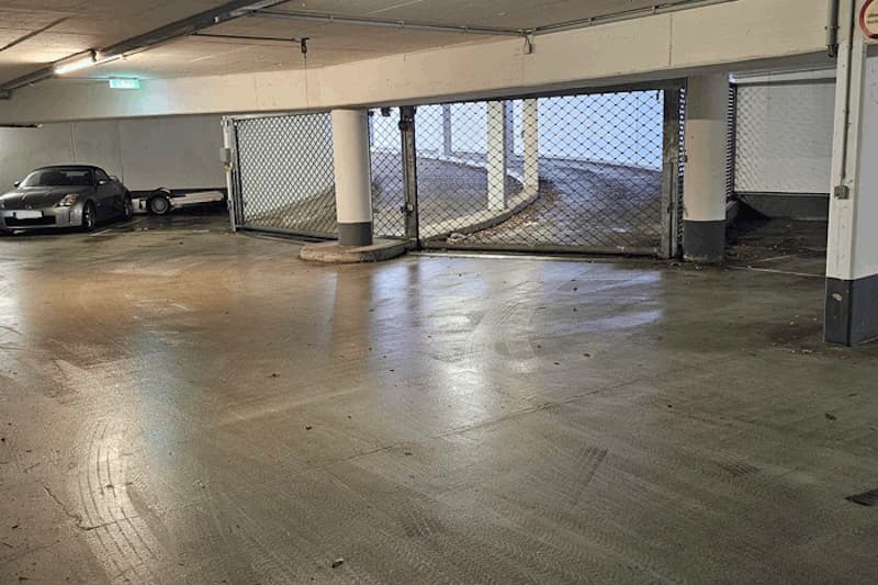 Leere Tiefgarage mit geschlossener Rollgittertür, Ausfahrtrampe, Säule und geparktem Auto mit Anhänger.