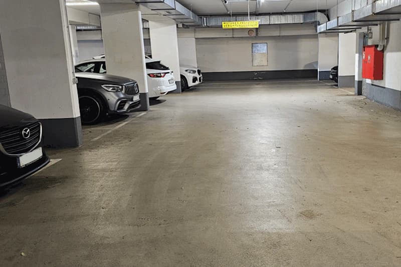 Tiefgarage mit Parkplätzen, Betonstützen, mehreren geparkten Autos links und Feuerlöscherkasten rechts.