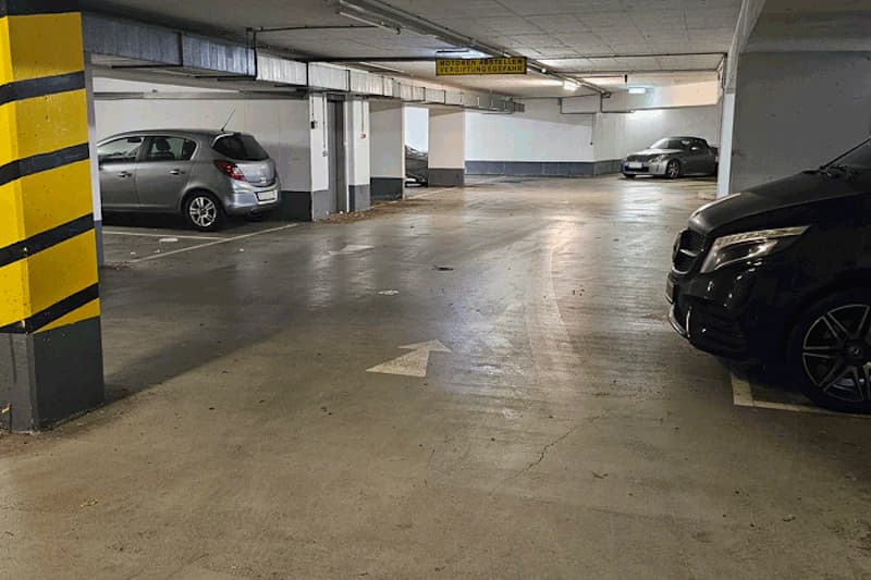 Tiefgarage mit mehreren geparkten Autos, Betonstützen mit gelb-schwarzen Streifen und Richtungspfeil auf dem Boden.