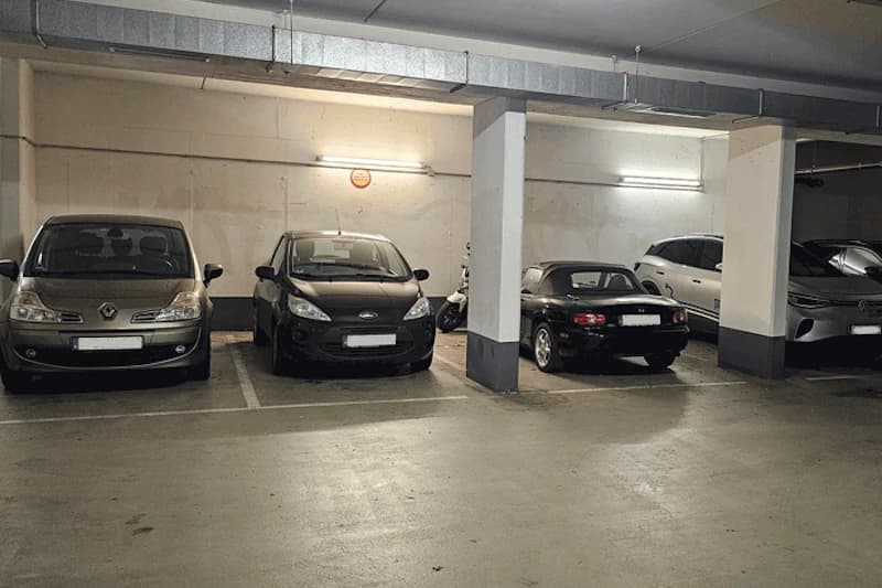 Tiefgarage mit fünf parkenden Autos, Betonpfeiler und Deckenleuchten, Motorroller zwischen zwei Fahrzeugen.