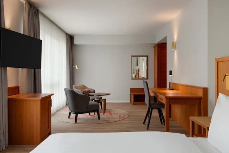 Schlafbereich mit Bett, Schreibtisch, zwei Sesseln, Kaffeetisch und Wandfernseher vor bodentiefem Vorhang.