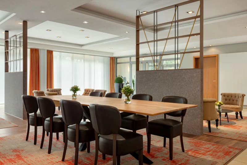 Langer Holztisch mit mehreren schwarzen Stühlen, Vasen mit Blumen, Trennwände und Sessel in hellem Hotelraum.