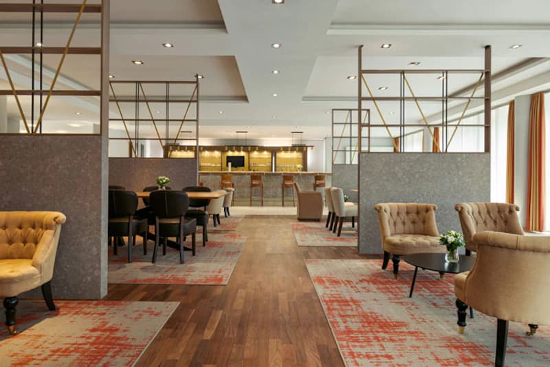 Hotel-Lounge mit Bar im Hintergrund, Polstersessel, Tische, Teppiche und Raumteiler, hell beleuchtet.