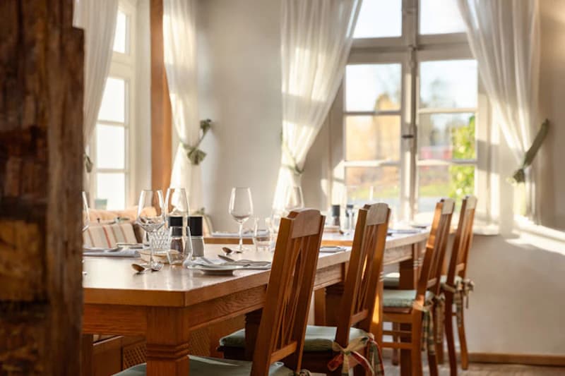 Sonnendurchfluteter Speisetisch mit Holzstühlen, eingedeckt mit Gläsern, Besteck und Salz- und Pfefferstreuern vor Fenstern mit Gardinen.