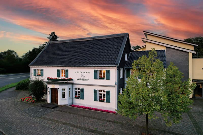 Weißes zweigeschossiges Haus mit grünen Fensterläden, Blumenbänken und beleuchteten Fenstern vor farbigem Abendhimmel.