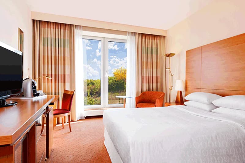Hell eingerichtetes Hotelzimmer mit großem Bett, Holzkopfteil, Schreibtisch mit Fernseher und Kaffeemaschine.