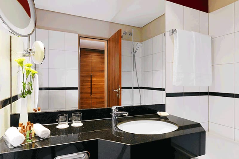 Badezimmer mit schwarzer Waschplatte, rundem Waschbecken, Spiegel, Handtuch, Duschbereich und offener Holztür.