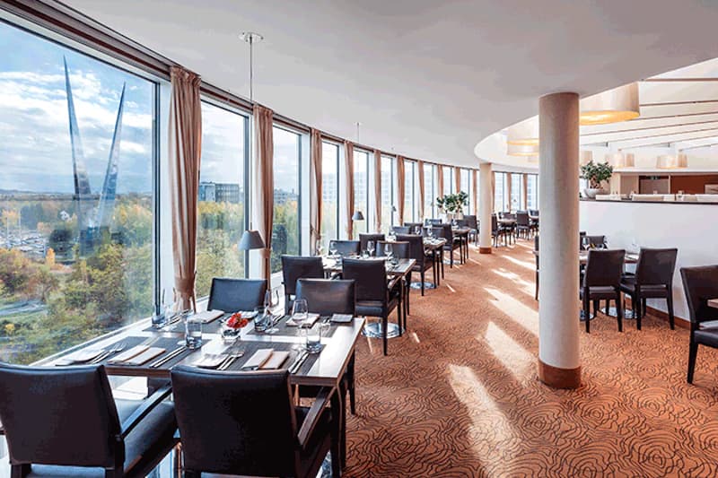Panorama-Restaurant mit gedeckten Tischen am Fenster und Blick auf Außenanlagen am Flughafen Düsseldorf.