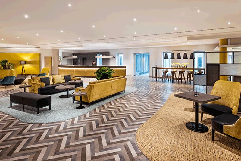 Hotelhalle mit Rezeption, gelben Sofas, Sesseln, Tischen, Pflanzen und gemustertem gemustertem Boden.