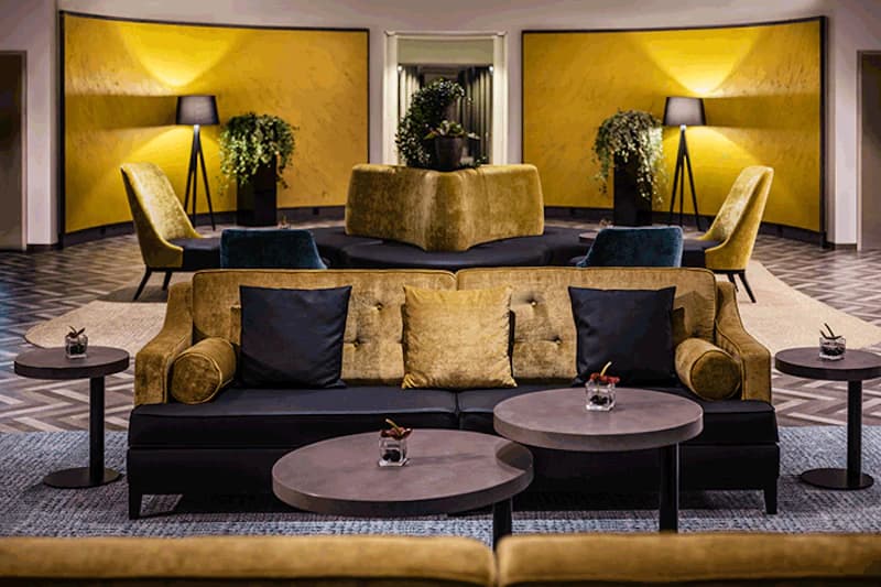Hotellounge mit Sofas, runden Tischen, Stehlampen und gelben Wandpaneelen.