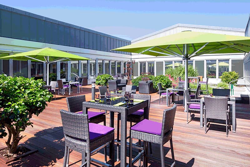 Außenterrasse mit Holzdeck, Rattanstühlen mit violetten Sitzkissen, grünen Sonnenschirmen und Glasfront.