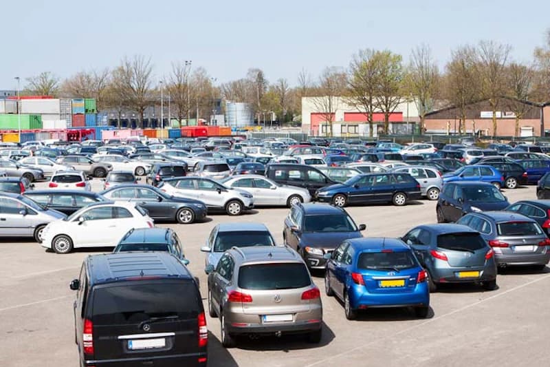 Großer gepflasterter Parkplatz mit vielen abgestellten Autos, im Hintergrund Container, Gebäude und Bäume.