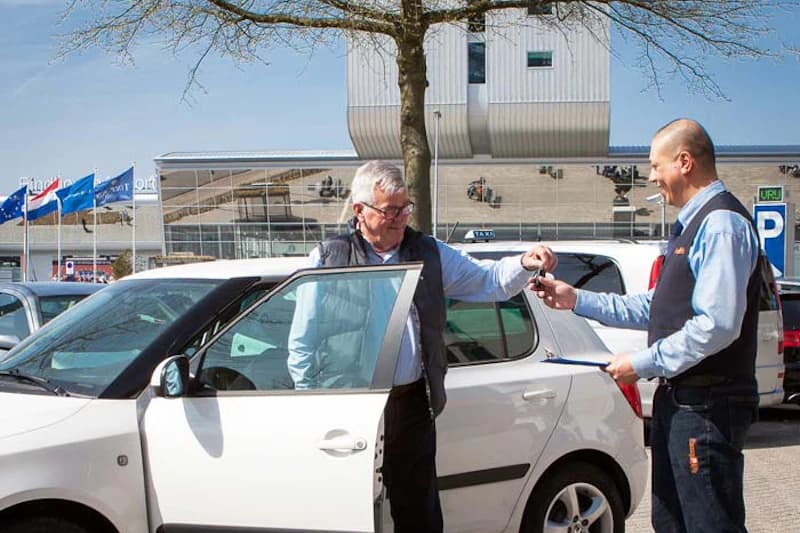 Älterer Mann erhält Autoschlüssel von Parkplatzmitarbeiter neben weißem Auto vor Flughafenterminal.