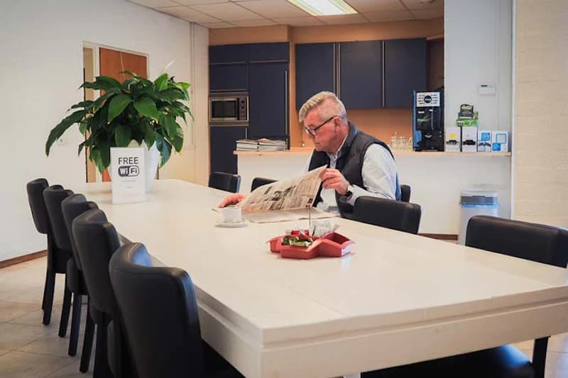 Mann liest Zeitung an langem weißem Tisch mit schwarzen Stühlen, Topfpflanze und Küchenküche im Hintergrund.